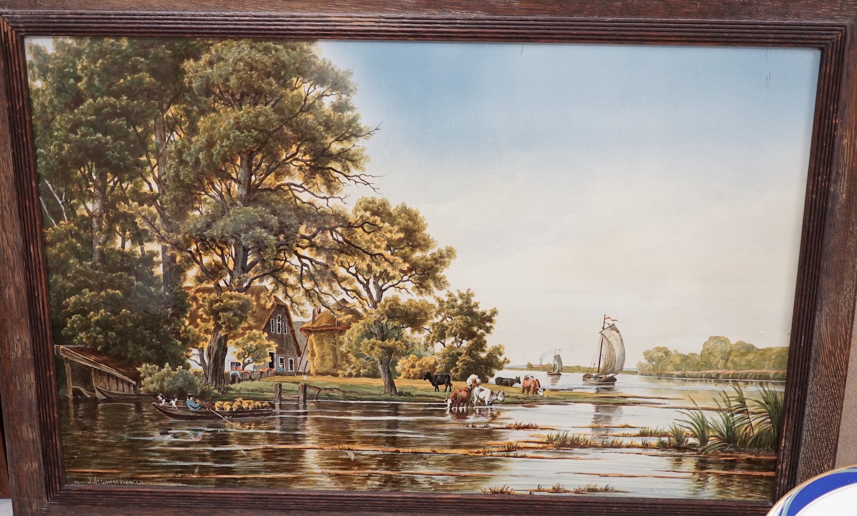 A framed continental ceramic plaque of a figurative country farm river scene, signed, 55cms wide x 35cms high